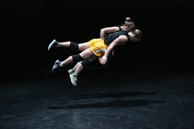 Two women are airborne parallel to the ground as if they were floating. Both women are wearing view is in profile. She wears a black tank top, orange athletic shorts, knee pads and sneakers.